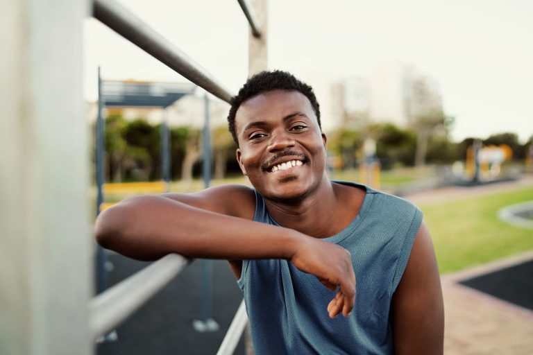 Smiling portrait of a sporty young african american man leaning on bar at the public park looking to camera with a big smile
