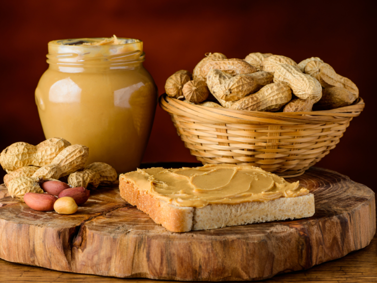 Jar of peanut butter with a bowl of peanuts and a piece of bread spread with peanut butter