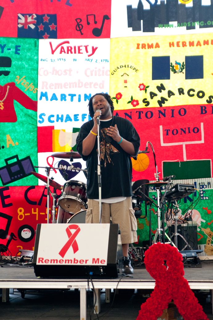 Man on stage singing into microphone with AIDS quilt panels behind him